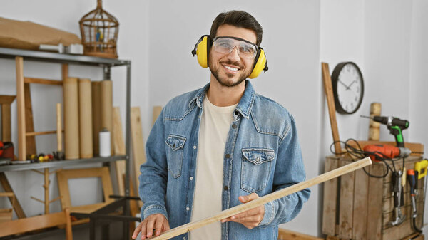 A smiling young hispanic man with a beard, wearing safety glasses and ear defenders, holds lumber in a well-equipped carpentry workshop.