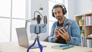 Kapalı mekanda mikrofon, kulaklık, akıllı telefon, dizüstü bilgisayar ve bitkilerle podcast yapan yakışıklı bir adam..
