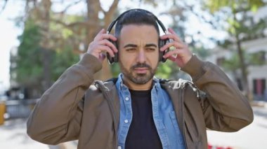 Sakallı bir İspanyol adam, parkın dışında beklerken kablosuz kulaklıkları ayarlıyor, eğlence ve teknolojiyi somutlaştırıyor..