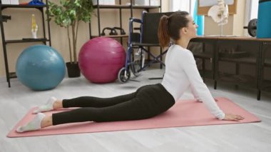Spor giyim bölümünde genç bir İspanyol kadın yoga yapıyor. İyi donanımlı bir spor salonunda pembe bir minderde poz veriyor.