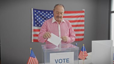 Orta yaşlı bir adam ABD bayraklarıyla içeride oy veriyor. Demokrasi ve vatanseverliği simgeliyor.