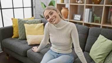 Şık bir ev dekoruyla rahat rahat oturma odasında oturan genç sarışın bir kadın..