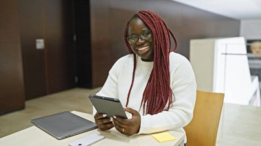 Saçları örgülü gülümseyen Afrikalı bir Amerikalı kadın modern bir ofiste tablet üzerinde çalışıyor.