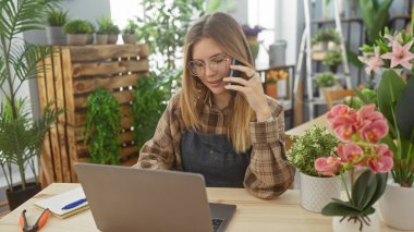 Genç bir kadın canlı bitkilerle çevrili bir çiçekçi dükkanında akıllı bir telefonla sohbet ediyor ve dizüstü bilgisayarı üzerinde çalışıyor..