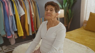 Kısa saçlı, olgun bir İspanyol kadın, rahat bir yatak odasında bir yatağın yanında oturur ve çeşitli giysiler giyer..