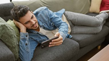Evdeki kanepede uzanmış gülümseyen genç İspanyol adam akıllı telefonuna günlük kıyafetlerle göz atıyor..