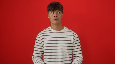 Çizgili tişörtlü yakışıklı bir İspanyol genç adam canlı kırmızı bir duvara karşı izole duruyor, ilginç bir ifadeyle hafifçe yana bakıyor.