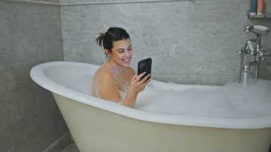 Neşeli bir kadın evde akıllı telefonunu evde kullanırken köpük banyosunda dinleniyor..