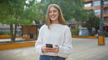 Şehir parkında akıllı telefon kullanan süveterli bir kadın.