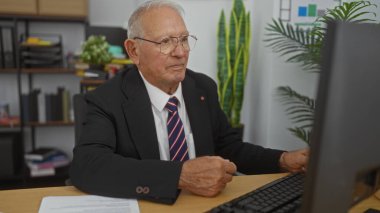 Ofis masasında çalışan üst düzey bir beyaz adam takım elbiseli ve gözlüklü kravat takıyor. Modern bir çalışma alanının ortasında odaklanmış ve profesyonel görünüyor..