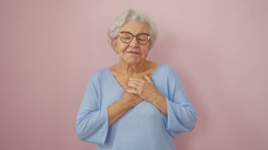 Gri saçlı, gözlüklü, pembe arka planda elleri kalbi olan sakin, yaşlı bir kadın, yumuşak bir duyguyu tasvir ediyor..