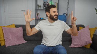 Neşeli bir adam, arka planda rahat bir oturma odası dekoru ve günlük kıyafetleriyle altı parmağını içeride sergiliyor..