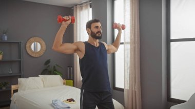 Yatak odasında sakallı bir adam halterleri kaldırıyor, spor ve sağlığa odaklanıyor..