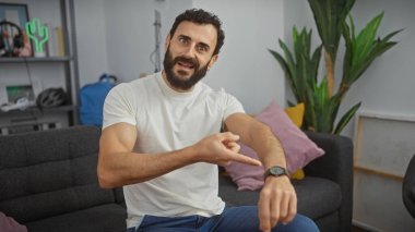 Modern oturma odasında oturmuş kol saatini gösteren sakallı, gülümseyen bir adam.