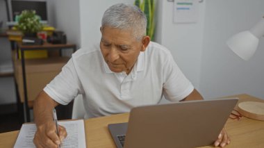 Beyaz saçlı İspanyol bir adam ofis ortamında not alıyor, bir masada dizüstü bilgisayarla oturuyor, modern, kapalı bir iş yerinde..