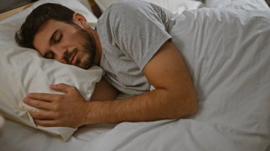 Genç İspanyol bir adam yatak odasında huzur içinde uyur, rahatlama, yaşam tarzı, rahatlık, yatak, ev, iç mekan, erkek, dinlenme ve refahı gösterir..