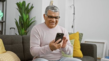 Beyaz saçlı, yakışıklı, olgun bir adam modern bir oturma odasında akıllı telefon kullanıyor..