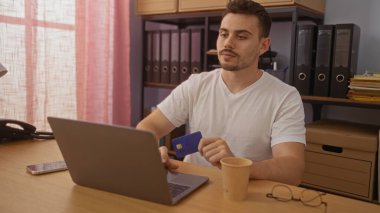 Elinde kredi kartı tutarken ve kahve içerken bilgisayarda çalışan sakallı yakışıklı İspanyol bir adam..