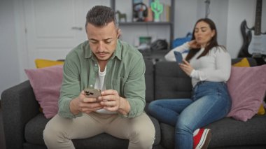 Odaklanmış bir kadın ve erkek, gündelik giysiler içinde, bağımsız ama birlikte, akıllı telefonlarıyla çağdaş bir oturma odasında buluşuyorlar..