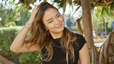 Radiantly confident young hispanic woman joyfully combing hair by hand in a green park, casting a cheerful smile to the side while relishing the fun outdoors clipart