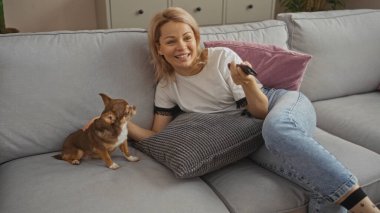 Sarışın genç bir kadın oturma odasındaki kanepede Chihuahua 'sıyla rahat bir şekilde ev ortamının tadını çıkarıyor..