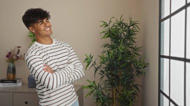 Yakışıklı, kollarını kavuşturmuş genç bir İspanyol adam aydınlık bir oturma odasında gülümsüyor, iç mekan bitkileri ve basit dekorlarla çevrili..