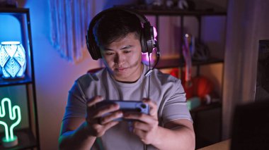 Genç Asyalı bir adam gece loş bir oyun odasında video oyunları oynuyor, akıllı telefonuna odaklanıyor..
