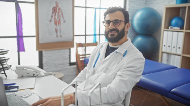 Emekçi kıyafetli olgun sakallı bir adam modern bir klinikte içten içe gülümsüyor..