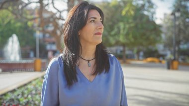 İspanyol olgun kadın park ortamında dalgın, güneş ışığı, ağaçlar, arka planda çeşme, şehir dışında açık alan.