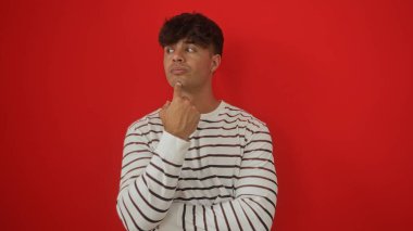 Kırmızı arka plan üzerinde izole edilmiş çizgili gömlekli yakışıklı İspanyol bir adam, düşünceli bir şekilde çenesine dokunuyor..