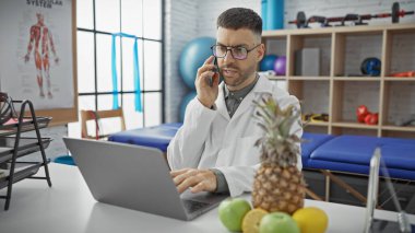 Laboratuvar önlüğü giymiş, parlak bir fizyoterapi kliniğinde telefon ve dizüstü bilgisayarla birden fazla iş yapan odaklanmış bir İspanyol..