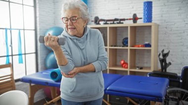 Hareket kabiliyeti ve sağlığı korumak için fizyoterapi kliniğinde dambılla egzersiz yapan yaşlı bir kadın..