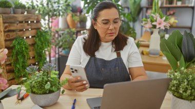 İspanyol kadın çiçekçide akıllı telefon kullanıyor. Etrafı bitkilerle çevrili, iş yerlerini dizüstü bilgisayarla ve bahçe aletleriyle düzenliyor..