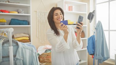 Parlak bir gardıropta kredi kartı tutarken akıllı telefonuyla selfie çeken neşeli bir kadın..