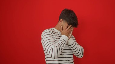 Yakışıklı İspanyol genç adam canlı kırmızı bir arka planda dururken elleriyle yüzünü gizler..