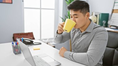 Modern ofis masasında kahve molası veren Asyalı genç bir adam. Etrafı temel işlerle çevrili.