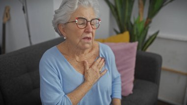 Gözlüklü yaşlı kadın oturma odası ortamında rahatsızlık yaşıyor, göğsüne dokunuyor..