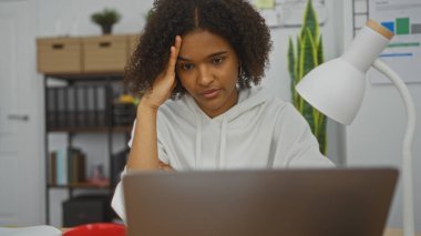 Kıvırcık saçlı Afro-Amerikalı genç bir kadın bilgisayarını iç ofis ortamında çalışırken odaklanmış görünüyor..