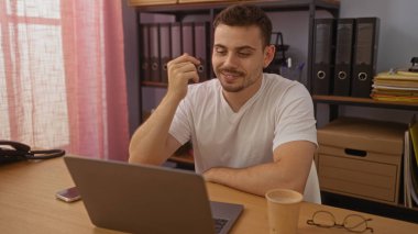 Ofiste elinde kahve fincanıyla dizüstü bilgisayarla çalışan yakışıklı İspanyol bir adam, profesyonel bir iç mekan ayarını yakalıyor..