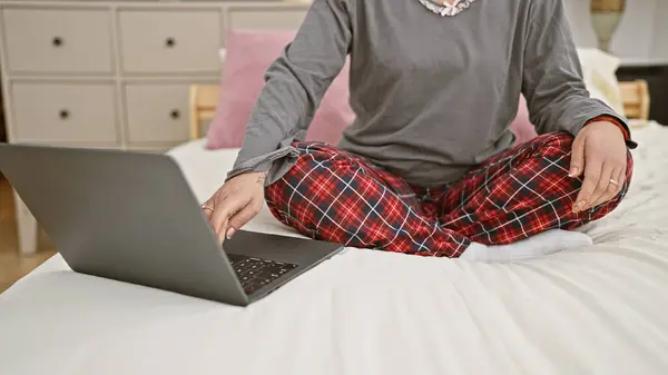 stock image An adult woman in pajamas works on a laptop while sitting cross-legged on a bed in a well-lit bedroom.