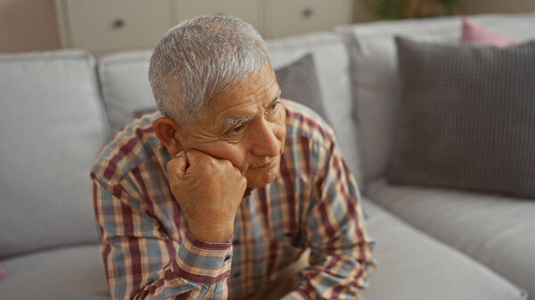 A thoughtful, grey-haired hispanic man sits in a living room, reflecting deeply indoors.