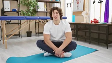 Genç, kıvırcık saçlı, sırıtan ve el kol hareketleriyle yoga minderinin üstünde oturan bir İspanyol..