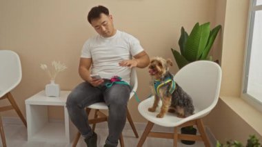 Klinikteki bekleme odasında genç bir adam köpeğini okşarken telefonunu kontrol ediyor, yanında beyaz bir sandalyede oturuyor..