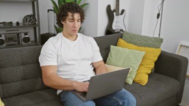 Evdeki kanepede oturan neşeli İspanyol genç adam, tamam işareti gösteren neşeli bir gülümseme sergiliyor, dizüstü bilgisayar keyfine dalmışken pozitif enerji yayıyor.