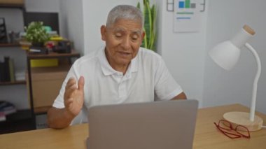 Ofis odasında çalışan, raflardan ve bitkilerden oluşan dizüstü bilgisayarıyla ilgilenen, olgun, gri saçlı, İspanyol bir adam.