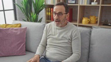Süveter giyen orta yaşlı İspanyol adam, evde şaşkın şaşkın oturuyor! Şaşırmış, ağzı tamamen açık, yüzünde korku resmedilmiş..