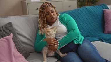Genç bir Afro-Amerikan kadın örgülü küçük köpeğini kucaklar rahat bir oturma odasında, yastıklarla çevrili bir dairede dinlenirken..
