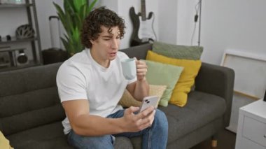 Kıvırcık saçlı genç bir adam rahat bir ev ortamında, elinde akıllı telefon ve kahve bardağıyla kanepede rahatlar..