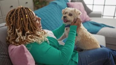 Saçları yeşil hırka giyen Afro-Amerikalı genç bir kadın renkli yastıkları ve büyük bir penceresi olan küçük köpeğini sevgi dolu bir oturma odasında tutuyor..