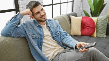 Yakışıklı İspanyol adam evdeki kanepede uzaktan kumandayla rahatlıyor.